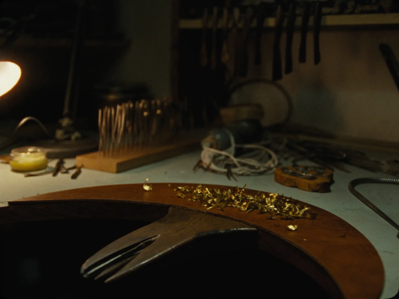 A jeweller's bench with a worn wooden bench pin, hand gravers in a rack, needle files, a ring clamp and brass shavings in the leather catch tray beneath.