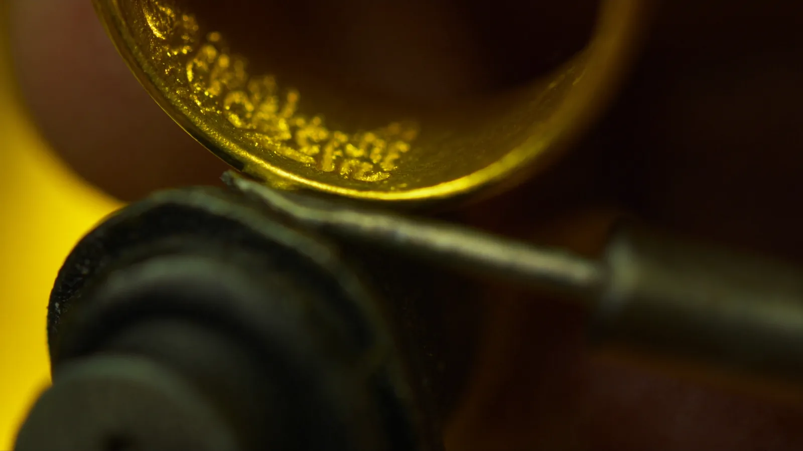 Macro view of a yellow gold band held in a ring clamp while script is cut into it by hand, with fine curls of gold raised by the graver.