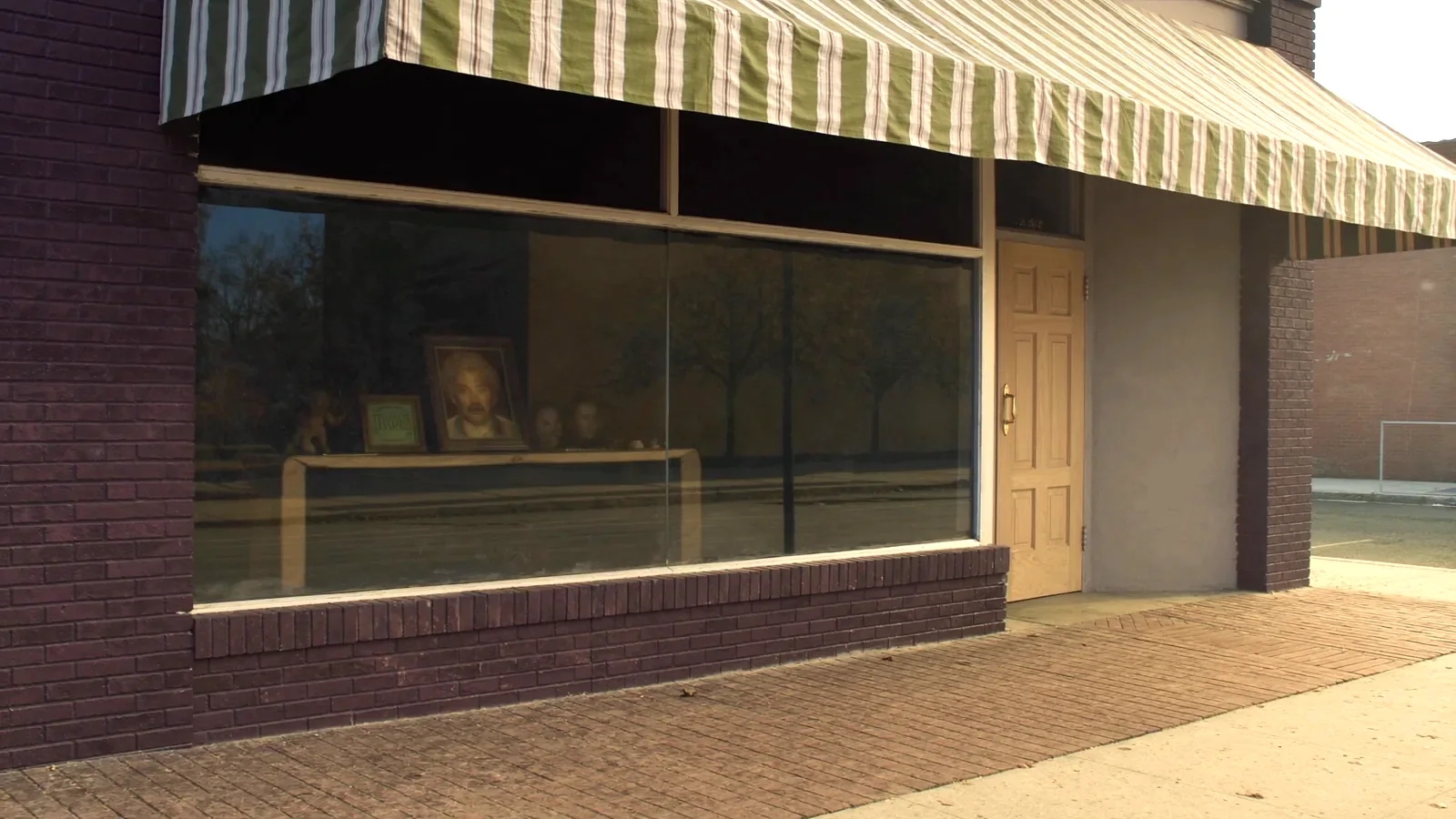 A narrow brick storefront with a large plate glass display window and a striped awning on a small town main street in autumn.